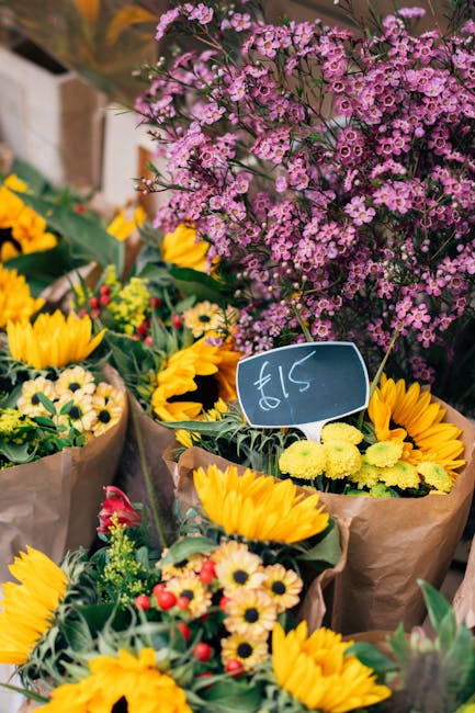 A vibrant display of fresh flower bouquets featuring bright yellow sunflowers, small yellow daisies, and red and white blossoms wrapped in brown paper, arranged on a table at an outdoor flower market.